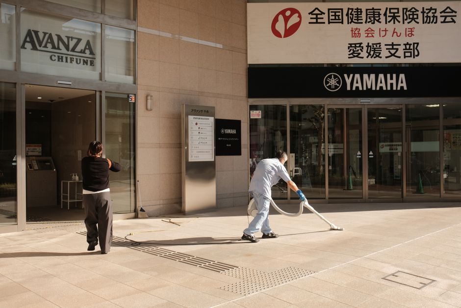 Two people clean the pavement outside a Yamaha building in Japan. Urban maintenance scene.