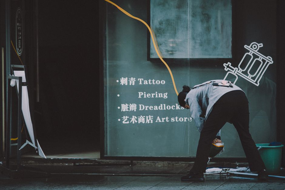 City worker cleans entrance of tattoo and piercing shop with visible signage.
